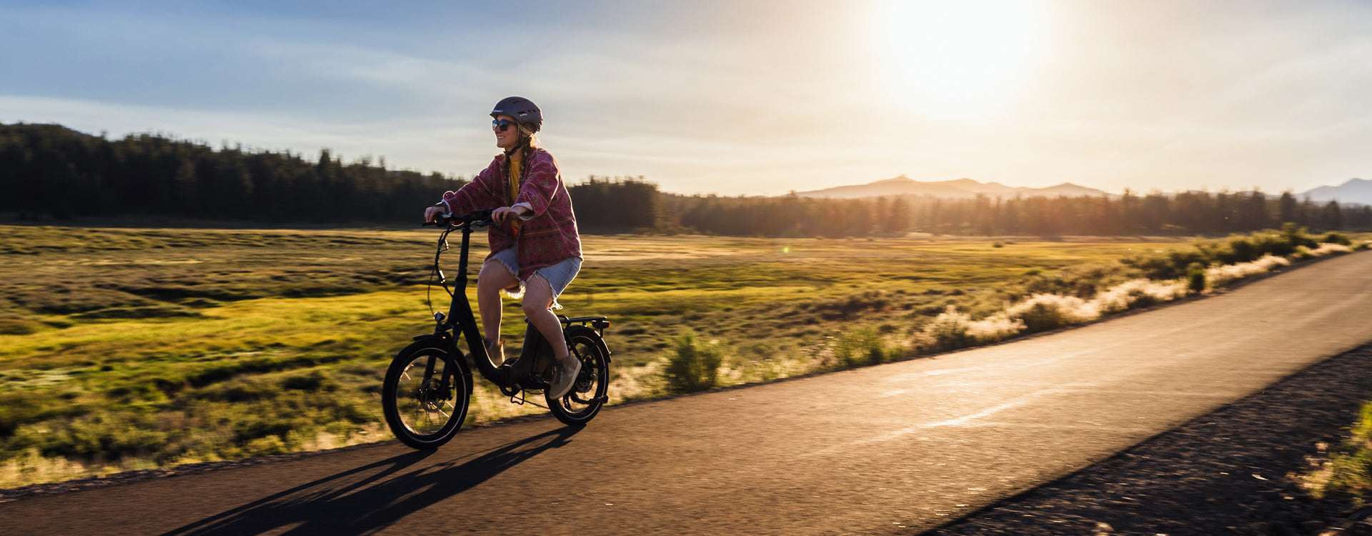 Woman riding an electric bicycle on a scenic path at sunset, surrounded by lush greenery and distant mountains.