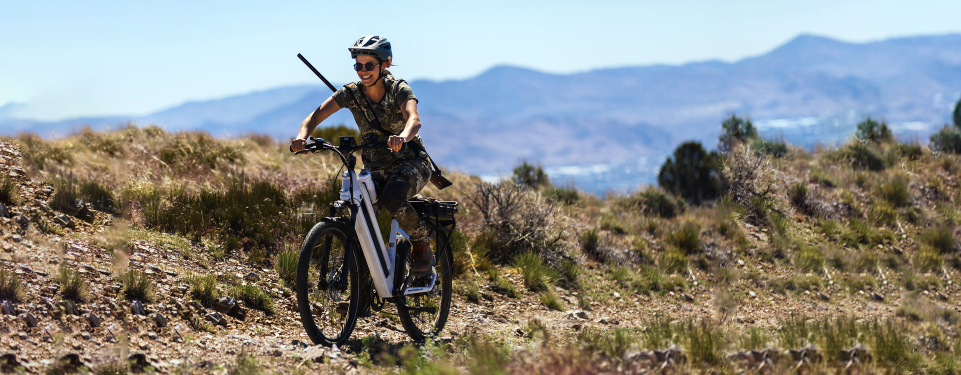 Person riding an electric bike in a mountainous terrain, wearing a helmet and camouflage outfit, with scenic hills and clear sky in the background.