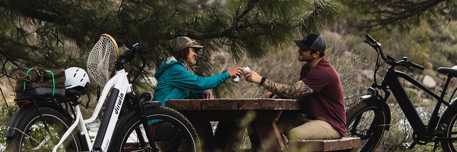 Two people enjoying a picnic at a wooden table in a forest setting, with electric bikes parked nearby, sharing drinks and smiling under tall pine trees.