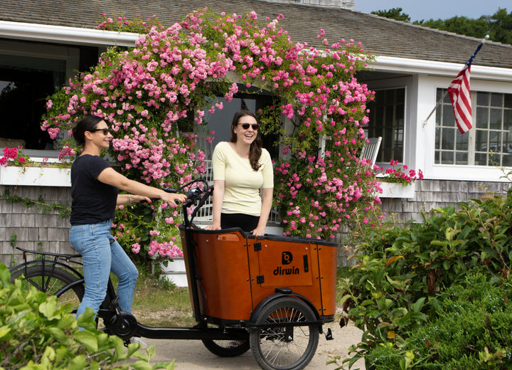 Two people with a cargo bike in front of a house with pink flowers.