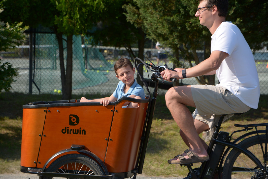 Man and child riding an orange Dirwin cargo bike in a park.