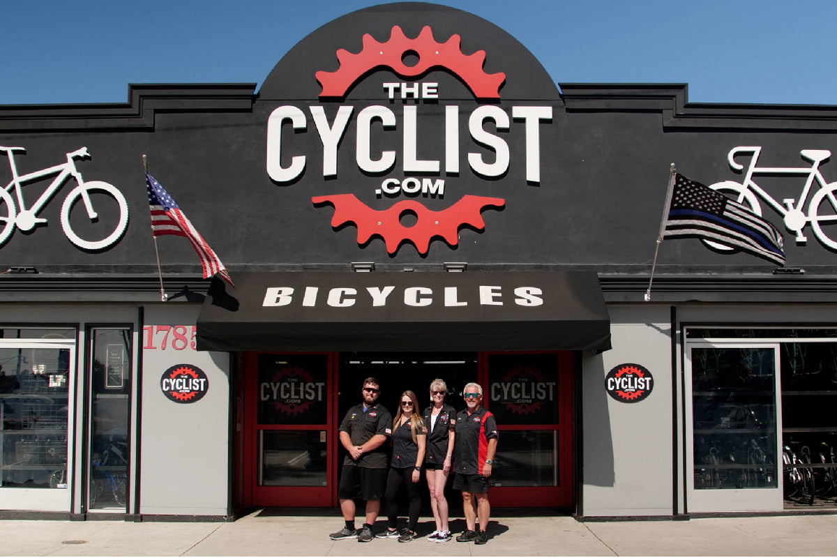 Exterior view of The Cyclist bike shop with four staff members posing in front, featuring prominent signage and American flags.